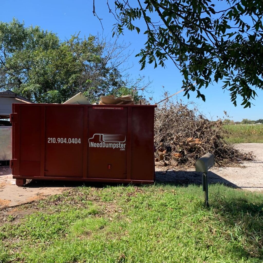 I Need Dumpster red dumpster sitting outside of a home that's having yard work done, large piles of tree branches are piled up beside the smaller dumpster