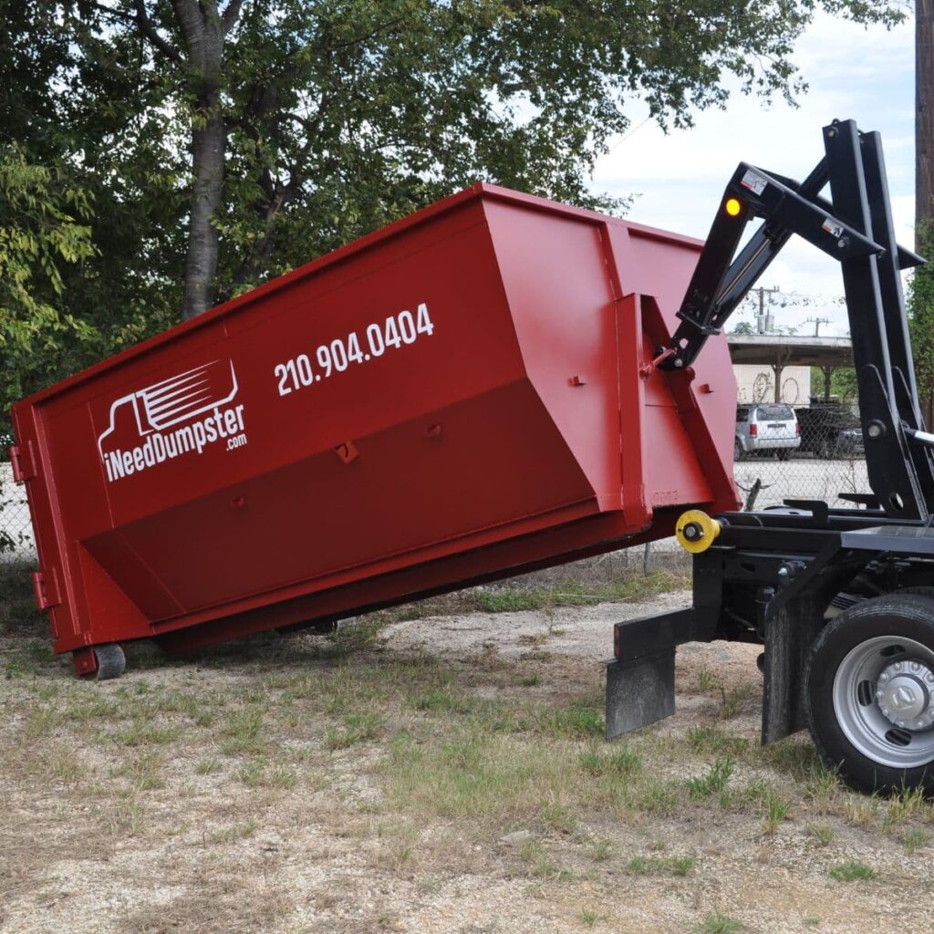 I Need Dumpster red dumpster being dropped off in a field next to a commercial area for a landscaping project