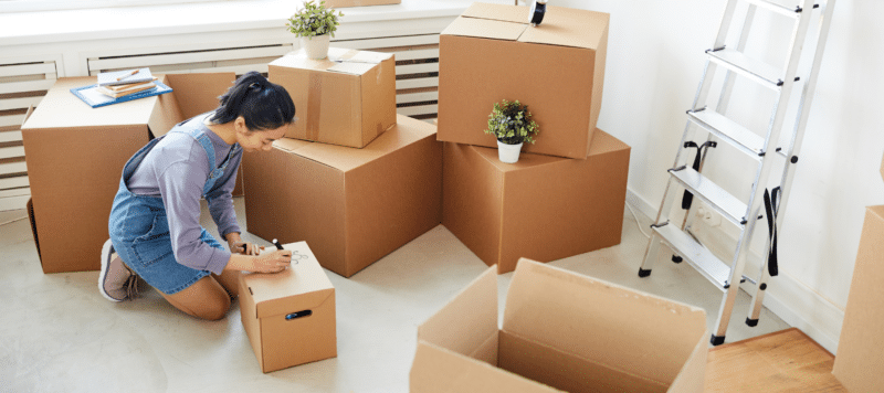 woman writing on a cardboard box surrounded by other cardboard boxes due to moving